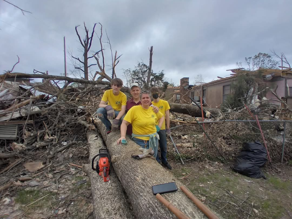 Family stands beside fallen tree after a tornado. They are wearing yellow clean-up-crew shirts. A chainsaw sits nearby. Behind them is a broken chainlink fence, six fallen trees and a house without a roof.